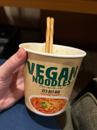 A paper cup with vegan ramen held in hand, two chopsticks sticking out